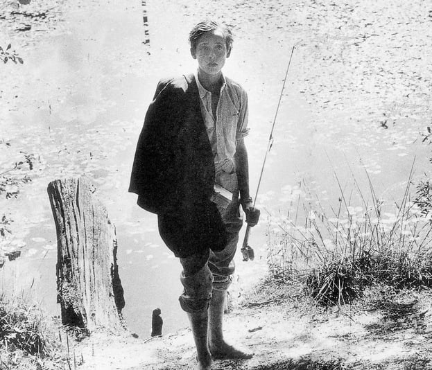 Black & white photograph of a teenage boy standing by a creek with fishing gear, looking devoid of emotion.