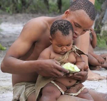 A Bushman sits on the ground and supports a child on his legs while holding food in front of the child to eat