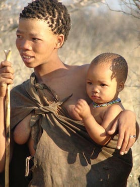 A Bushman mother standing holding an infant at her side slung in a wrap