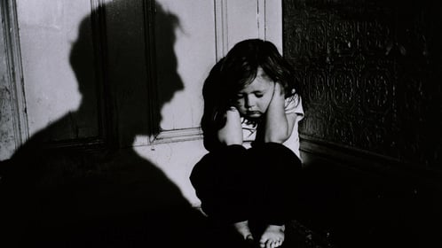 A young girl cowers in the corner with her hands over her ears with a silouette on the wall of a parent screams at her