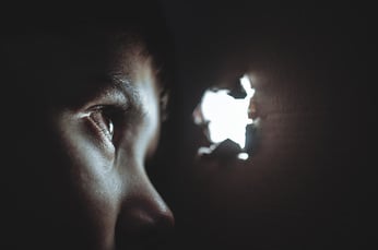 A young person in the dark looks through a small hole in the wall to the light on the outside