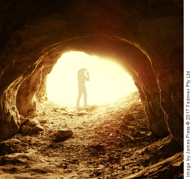 Looking from inside a dark cave a man stands at the exit shielding his eyes with his hands from the bright light outside