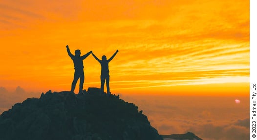 Two people stand on the summit with arms raised with a golden sun in the background