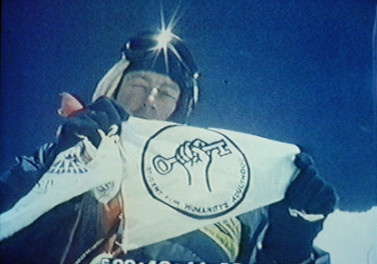 Tim Macartney-Snape standing on the summit of Mt Everest holding the FHA flag with the logo of a hand holding a key aloft.