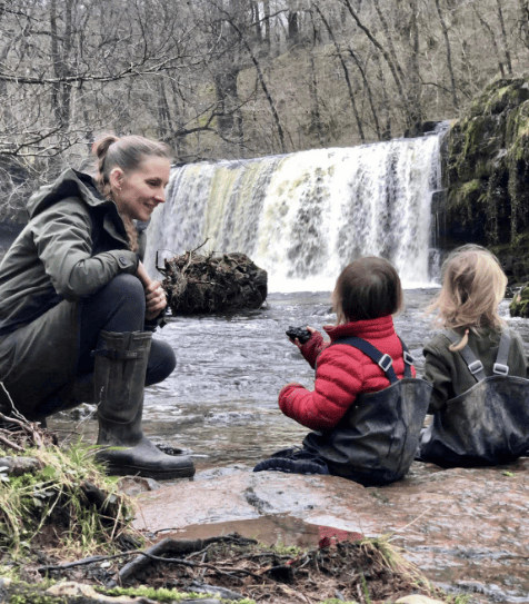 Beth smiling looking at 2 small children sitting on a rock beside a waterfall
