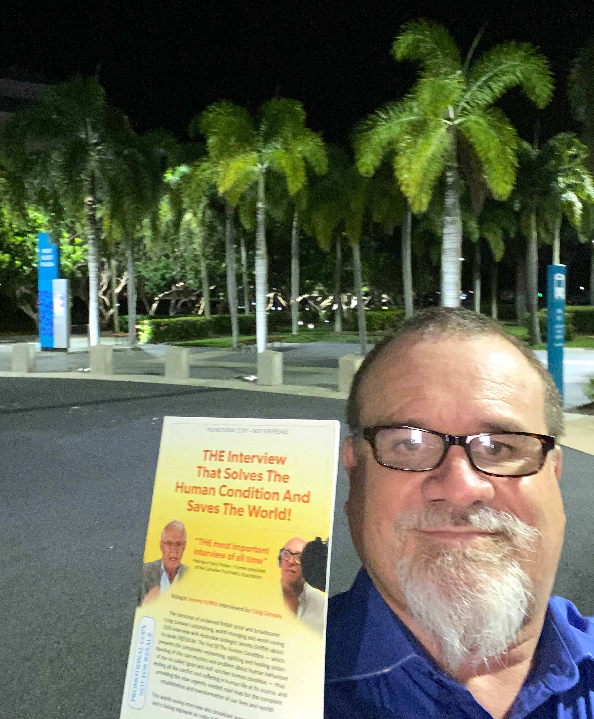 Joseph holding a copy of the book 'THE Interview' with palm trees lit up by lights in background
